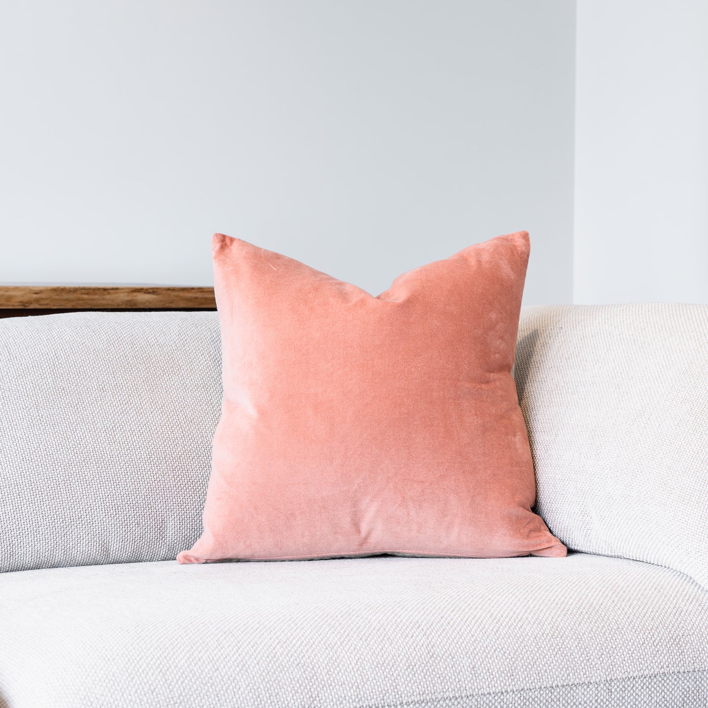 A square coral pink velvet throw pillow resting on a light gray textured fabric sofa against a neutral light gray wall.