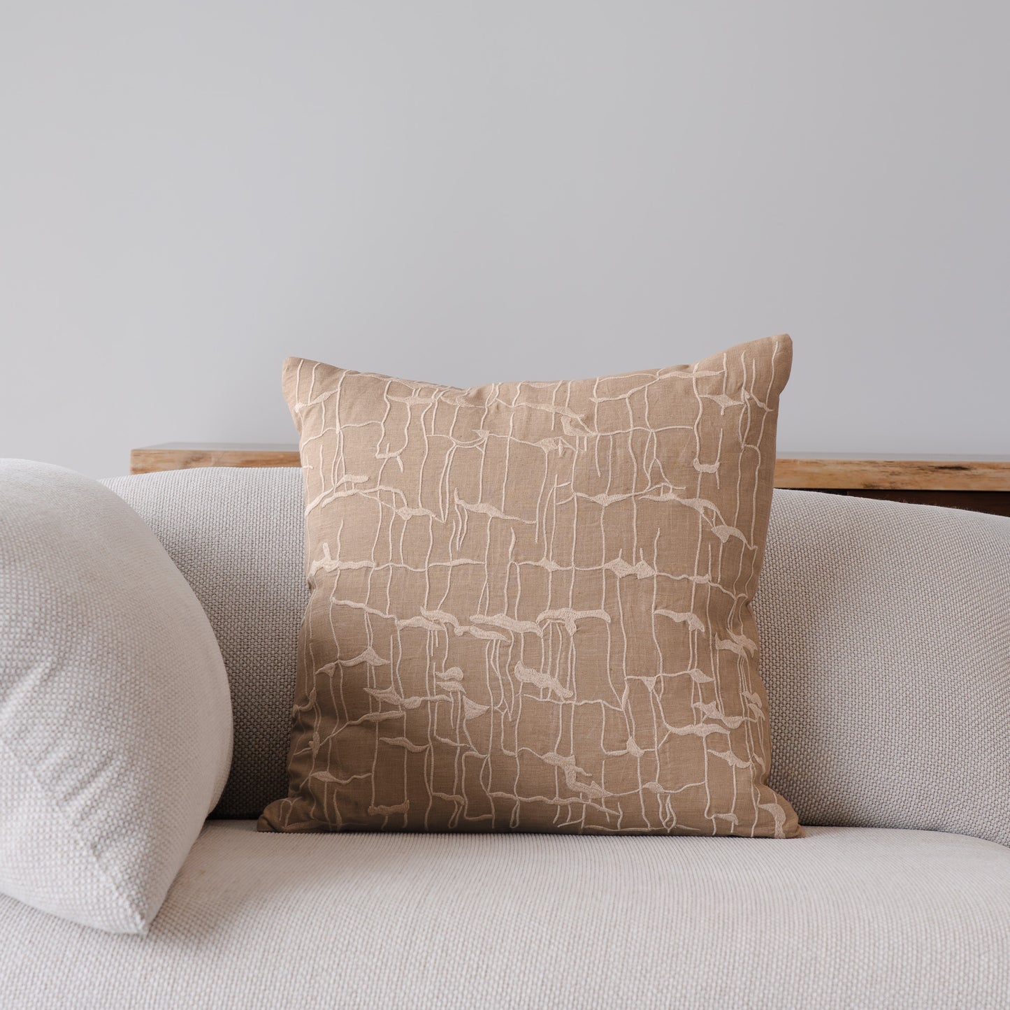 A square throw pillow in a warm beige fabric with a white abstract line pattern, styled on a light gray sofa next to another plain pillow.