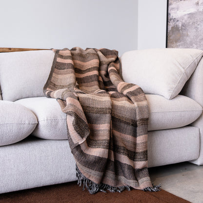 A striped woven throw blanket in shades of brown, beige, and muted pink is draped over a light gray fabric sofa in a modern living room setting.