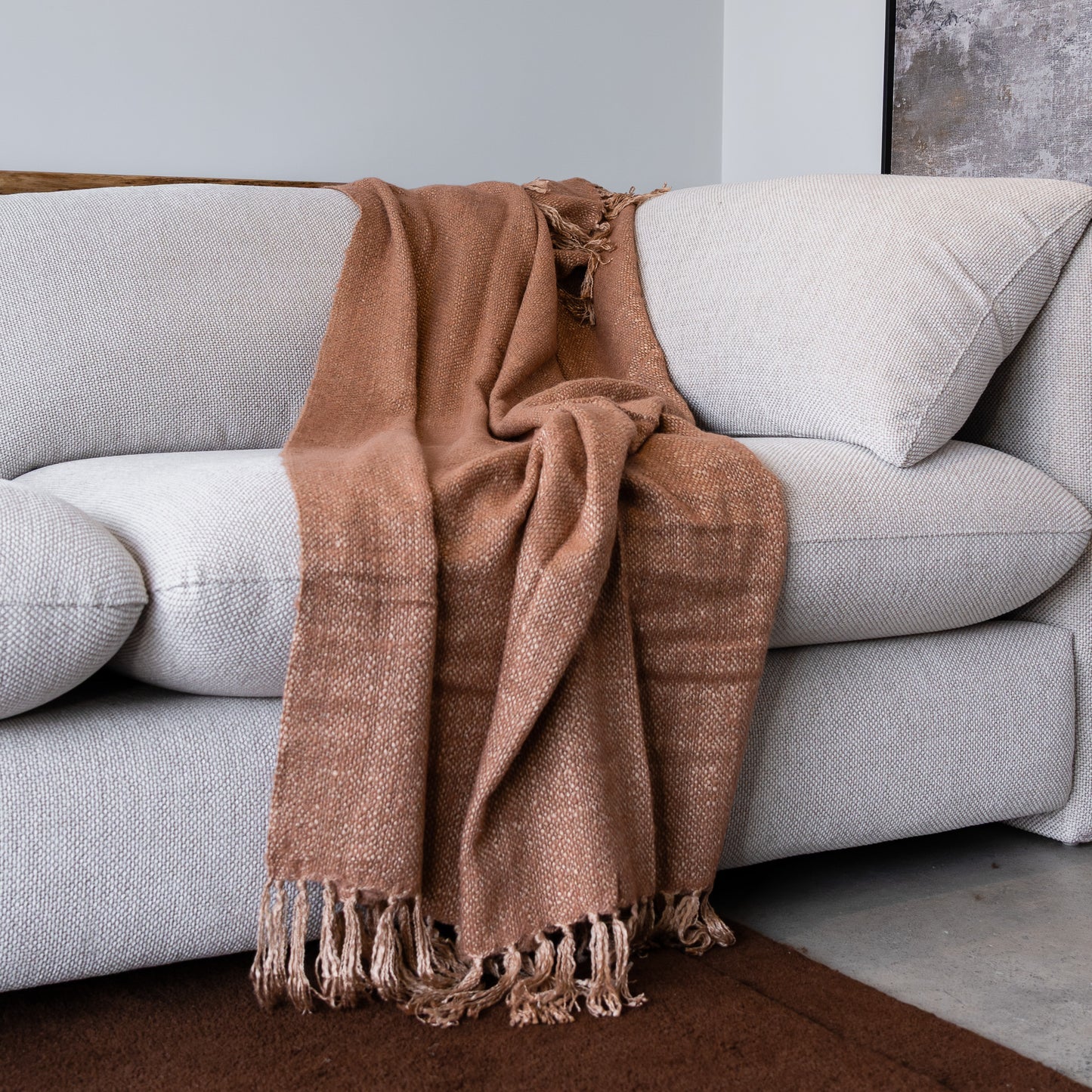 A warm brown woven throw blanket with fringe tassels draped over a light gray fabric sofa in a modern living room setting.