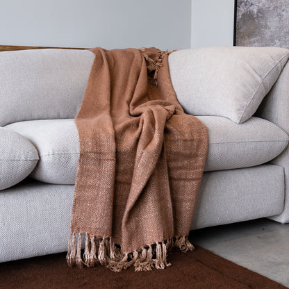A warm brown woven throw blanket with fringe tassels draped over a light gray fabric sofa in a modern living room setting.