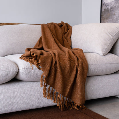 A warm brown woven throw blanket with fringe detailing, draped over a light gray textured sofa in a modern living room setting.