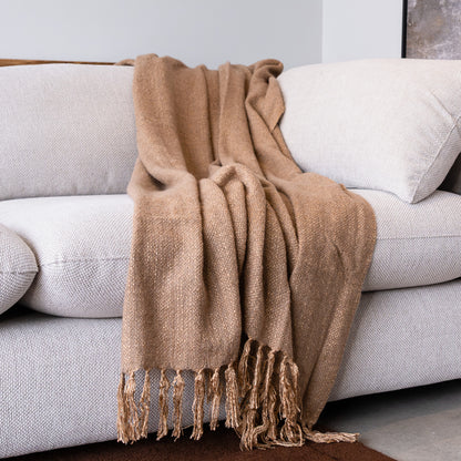 A warm camel-colored woven throw blanket with fringed edges draped over a light gray fabric sofa.