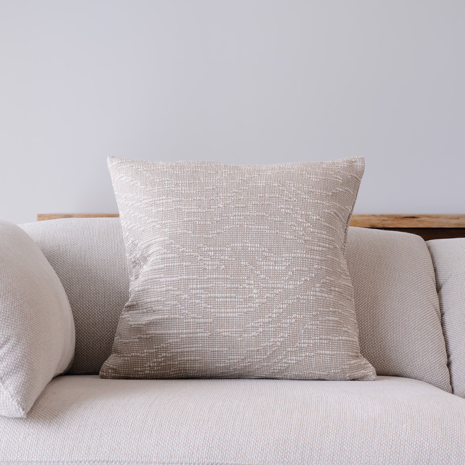 A square throw pillow with a textured, woven beige and light gray pattern, styled on a light gray fabric sofa against a neutral gray wall.