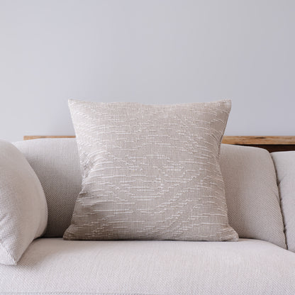 A square throw pillow with a textured, woven beige and light gray pattern, styled on a light gray fabric sofa against a neutral gray wall.