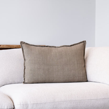 A rectangular lumbar throw pillow in a textured taupe linen fabric with a dark brown fringed edge, resting on a light beige textured sofa against a neutral gray wall.
