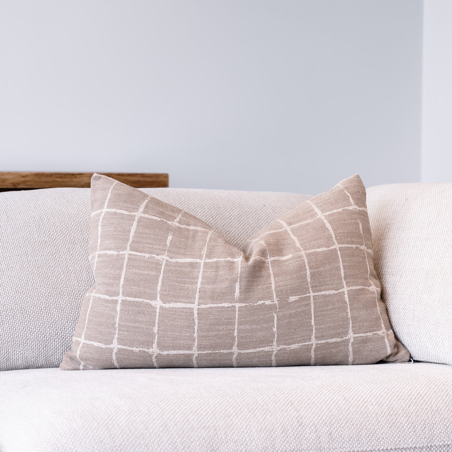 A square throw pillow with a beige and white grid pattern, resting on a light beige textured sofa against a neutral gray wall.