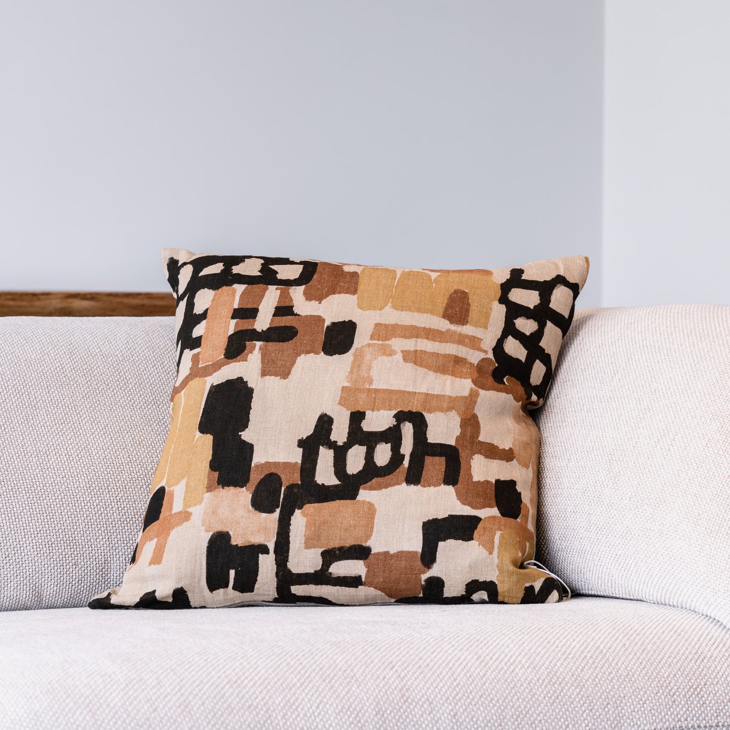 A square throw pillow with an abstract geometric pattern in beige, brown, and black, resting on a light gray textured sofa against a light gray wall.