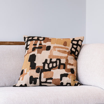 A square throw pillow with an abstract geometric pattern in beige, brown, and black, resting on a light gray textured sofa against a light gray wall.