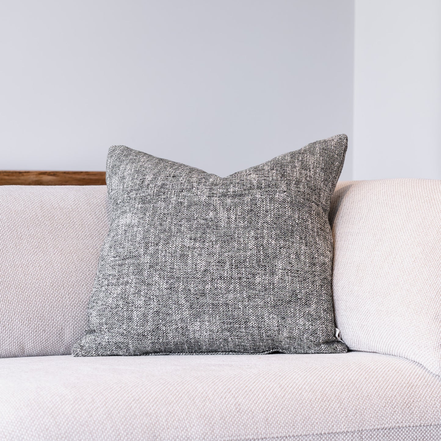A square gray textured throw pillow resting on a light beige sofa against a neutral gray wall.