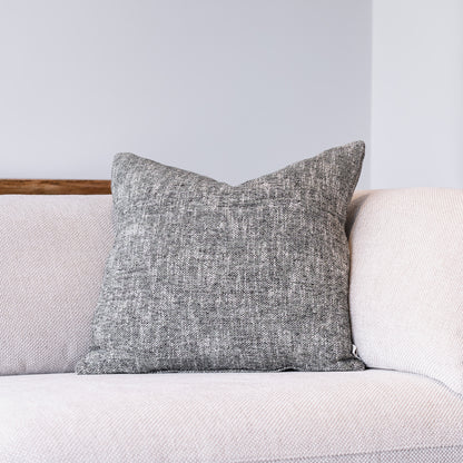A square gray textured throw pillow resting on a light beige sofa against a neutral gray wall.