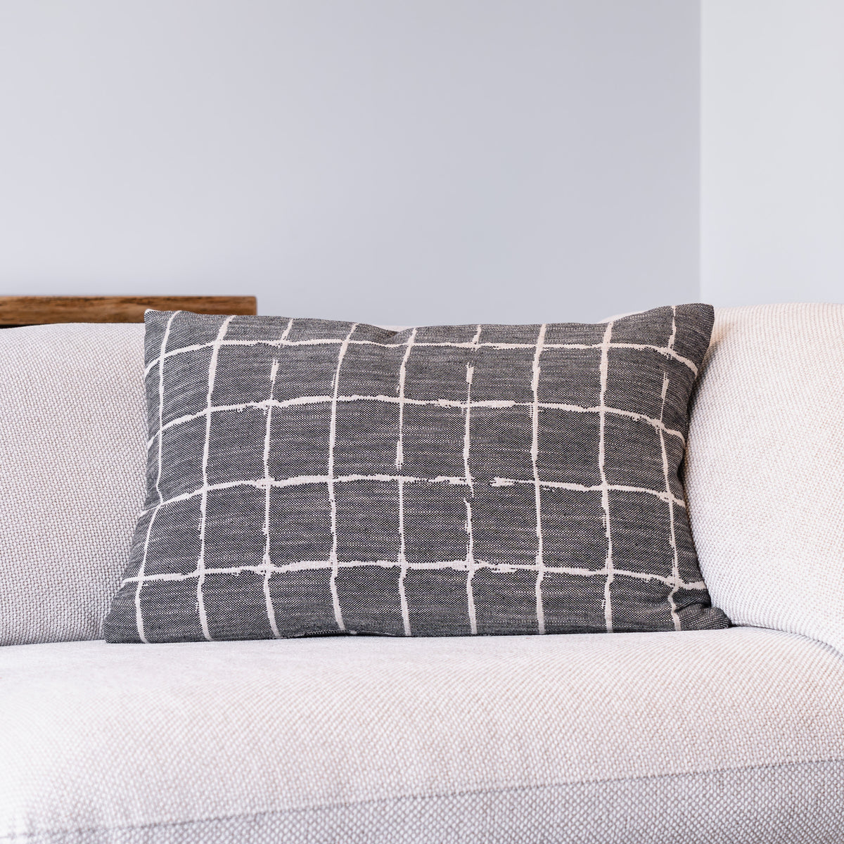 A rectangular throw pillow with a white grid pattern on a dark gray textured fabric, resting on a light beige textured sofa against a light gray wall.
