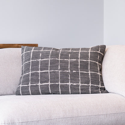 A rectangular throw pillow with a white grid pattern on a dark gray textured fabric, resting on a light beige textured sofa against a light gray wall.