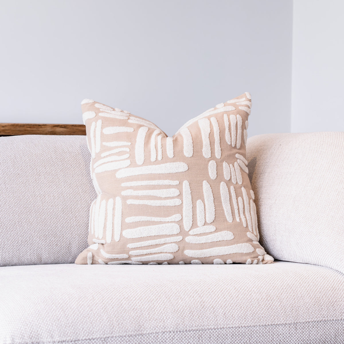 A square throw pillow with a beige and white abstract brushstroke pattern, resting on a light gray textured sofa against a neutral gray wall.