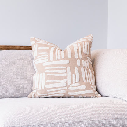A square throw pillow with a beige and white abstract brushstroke pattern, resting on a light gray textured sofa against a neutral gray wall.
