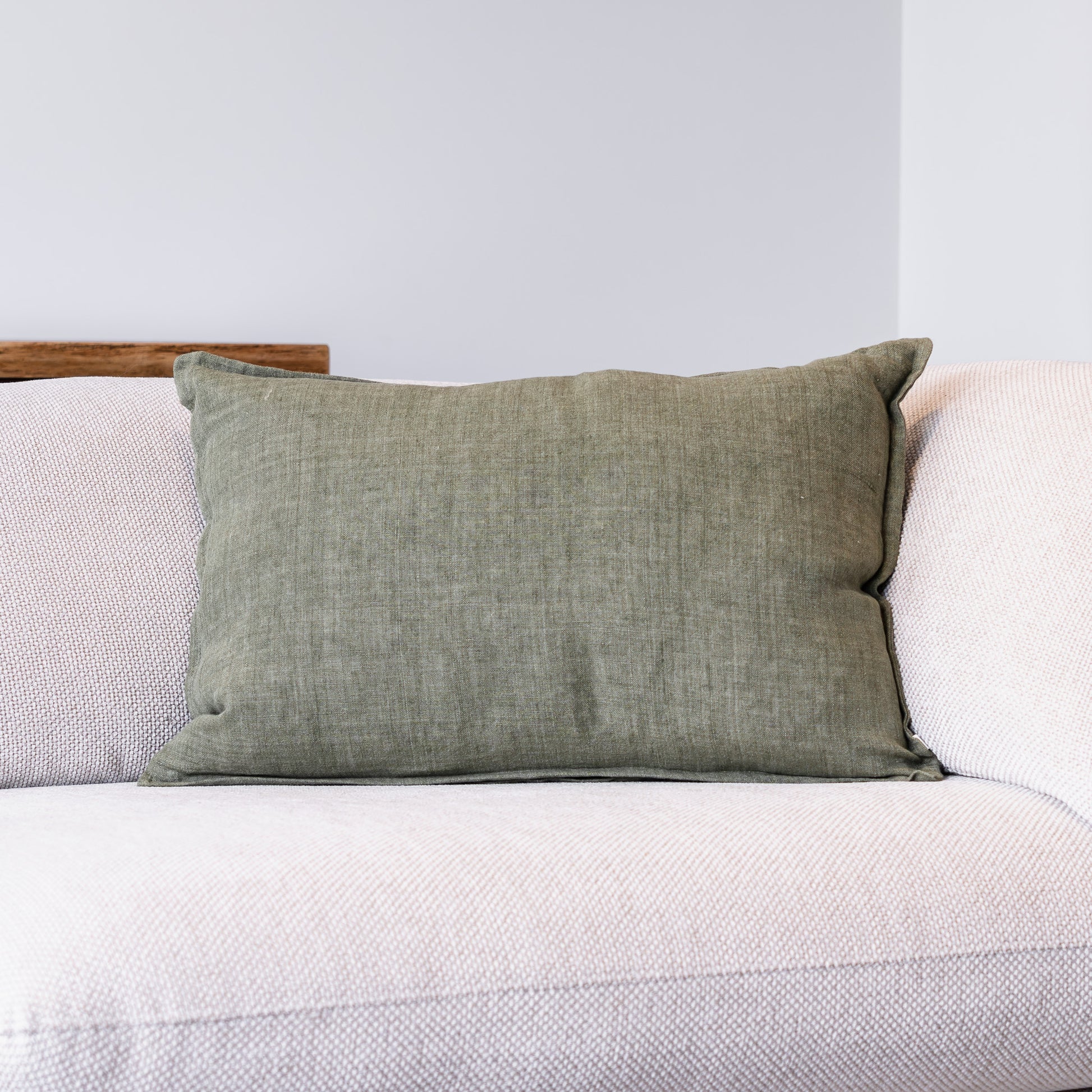 An olive green textured throw pillow resting on a light beige textured sofa against a neutral gray wall.