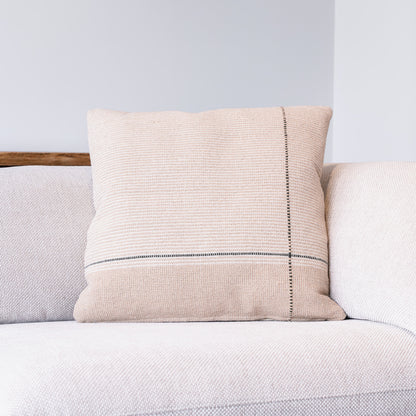 A square beige woven throw pillow with a vertical black stitched line and a horizontal black stripe, resting on a light-colored textured sofa.