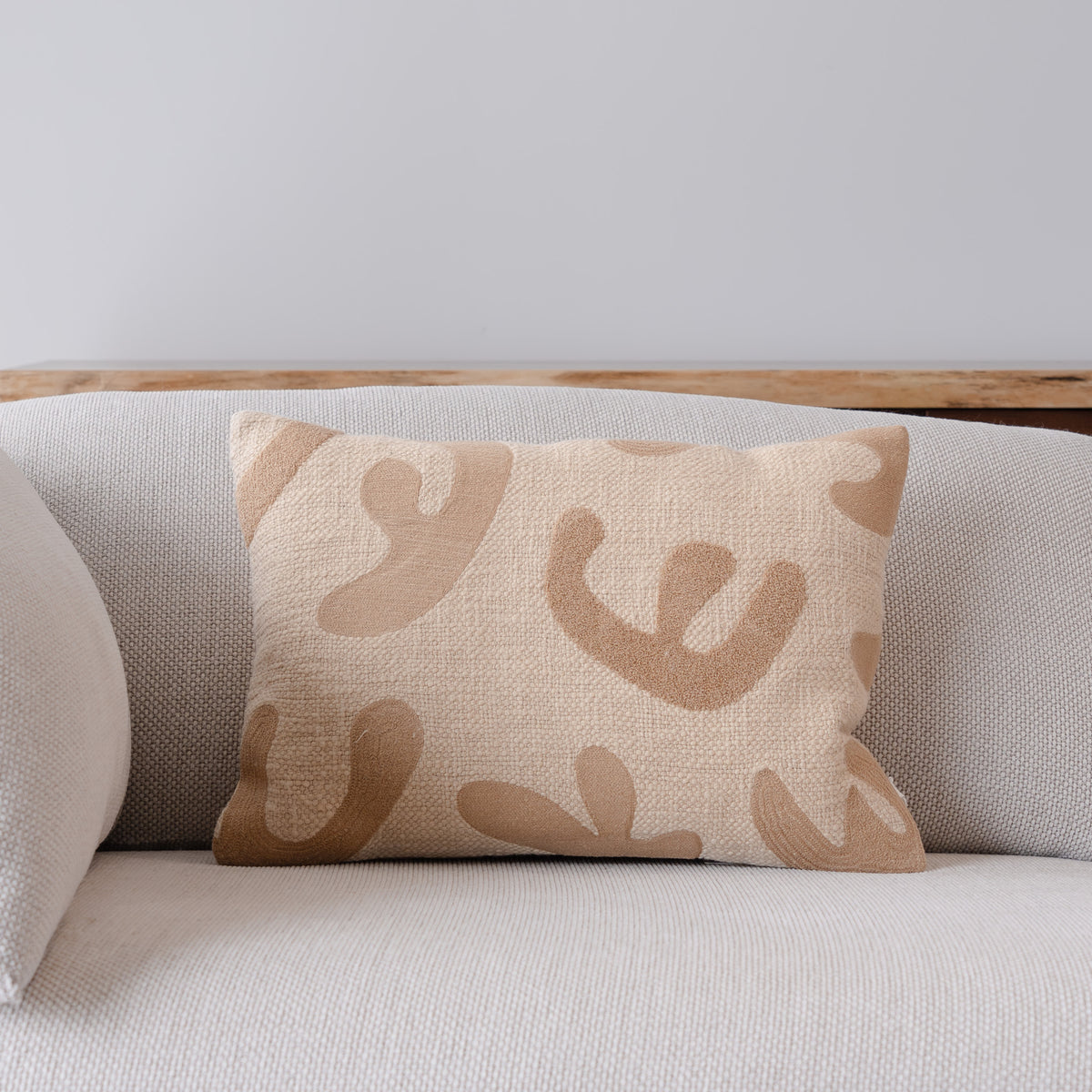 A rectangular throw pillow with a beige and tan abstract organic pattern, resting on a light beige textured sofa.