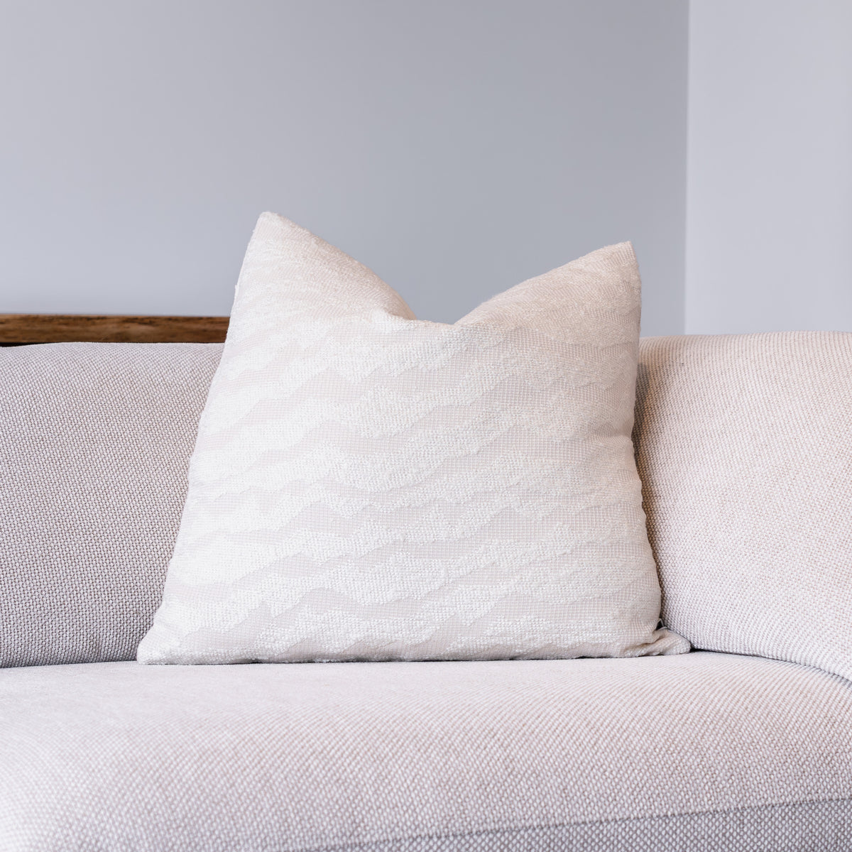 A square off-white throw pillow with a subtle wavy jacquard pattern, resting on a light beige textured sofa against a neutral gray wall.