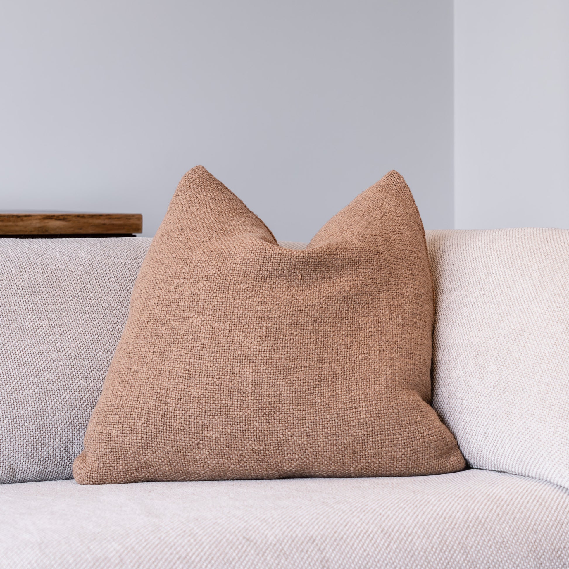 A square throw pillow in a textured tan linen fabric, styled on a light beige sofa against a neutral gray wall.