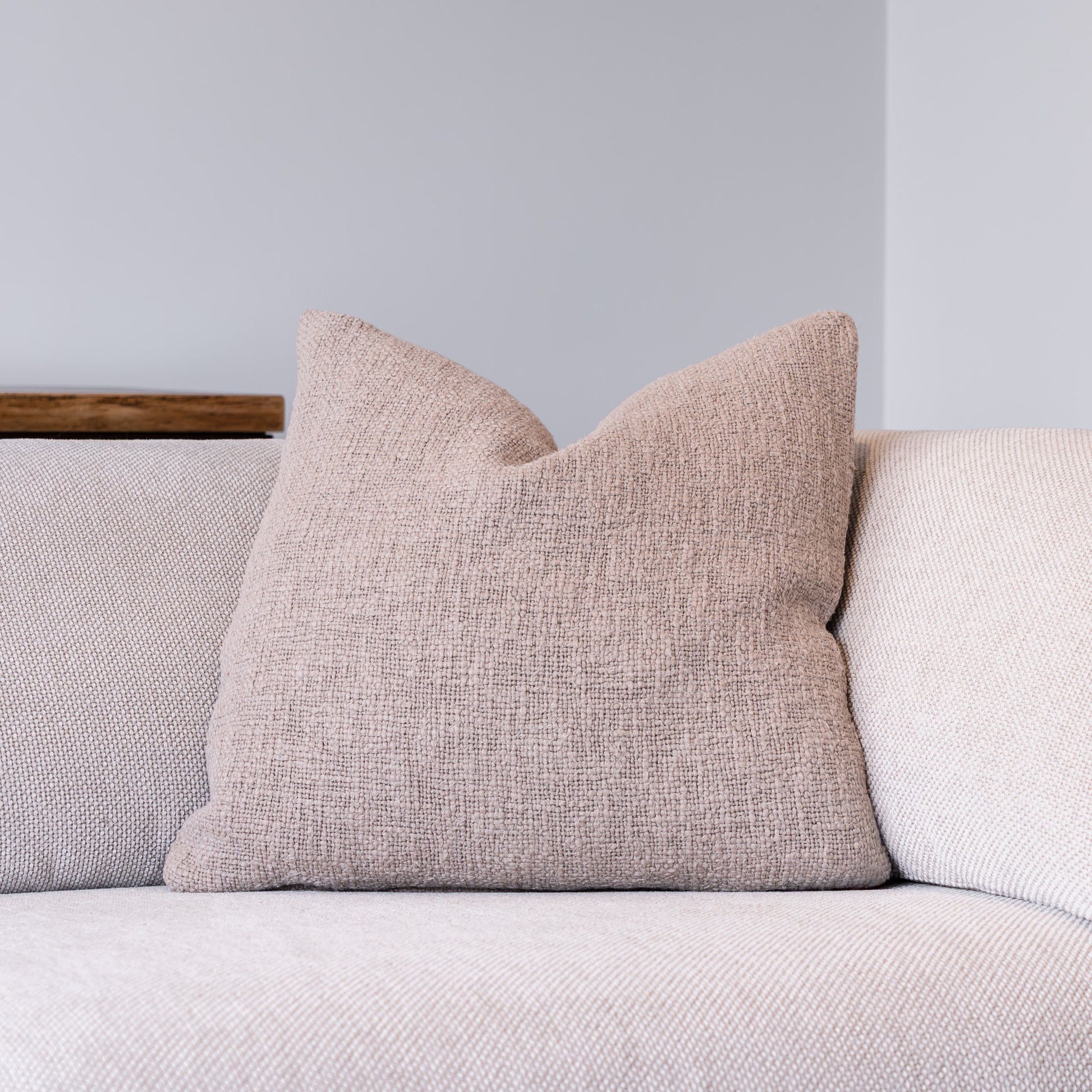 A square throw pillow in a textured, woven dusty pink fabric, resting on a light beige sofa against a neutral gray wall.