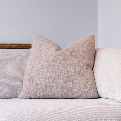 A square throw pillow in a textured, woven dusty pink fabric, resting on a light beige sofa against a neutral gray wall.