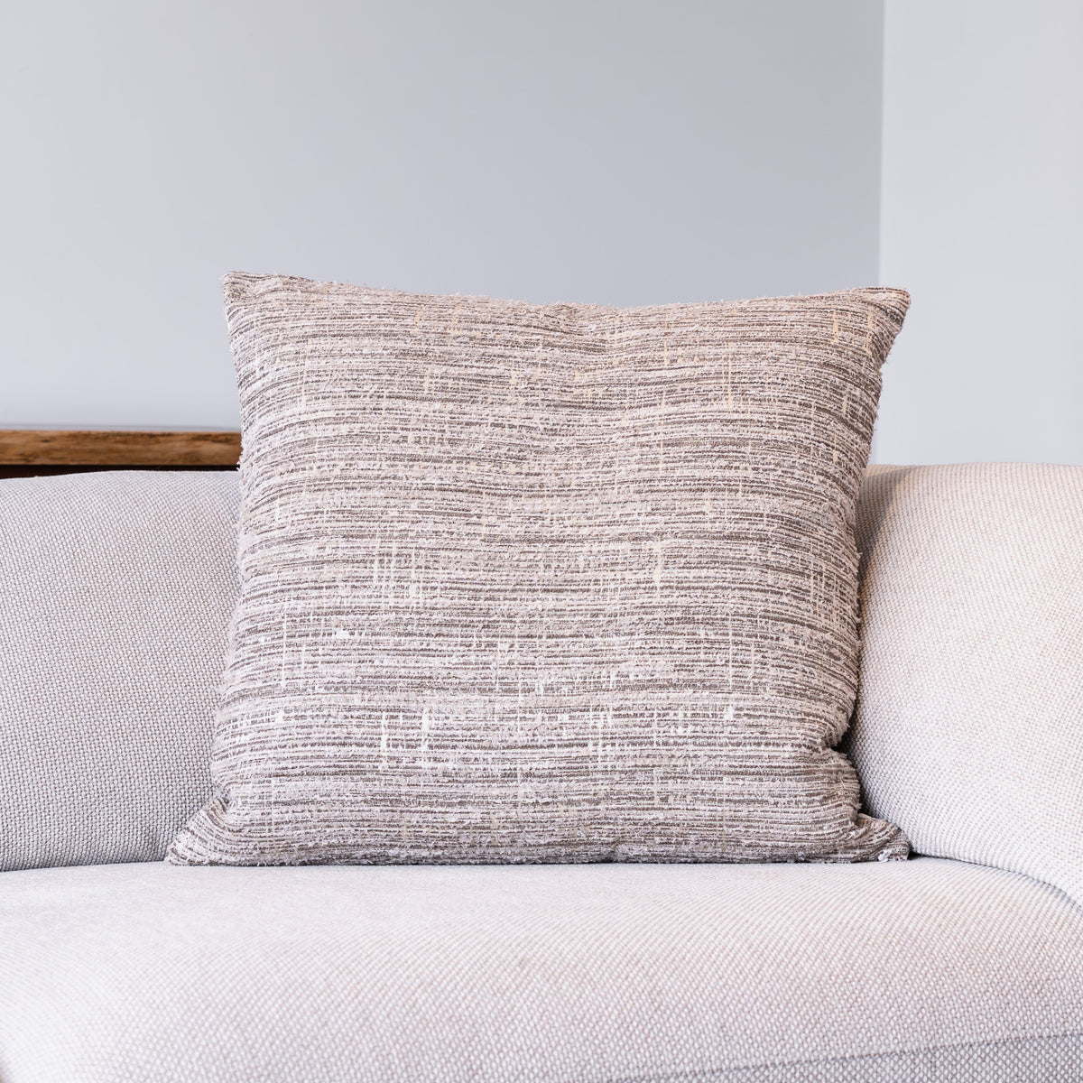 A square throw pillow with a textured, heathered taupe and beige fabric, resting on a light gray sofa against a neutral gray wall.
