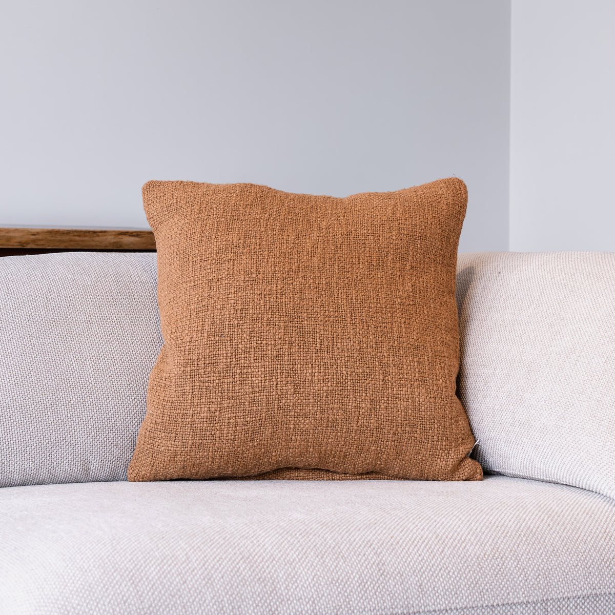 A square throw pillow in a textured rust-orange fabric, styled on a light gray textured sofa against a neutral gray wall.