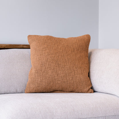 A square throw pillow in a textured rust-orange fabric, styled on a light gray textured sofa against a neutral gray wall.