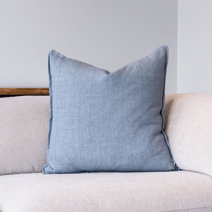 A square light blue throw pillow with piped edges, resting on a light beige textured sofa against a neutral gray wall.