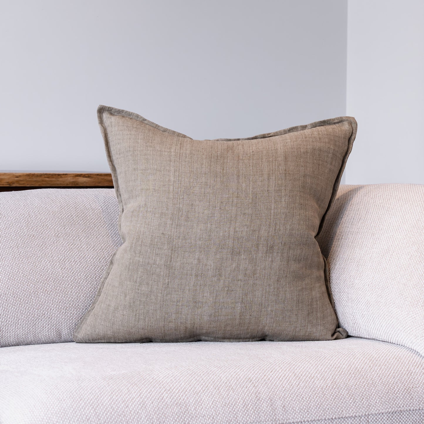 A square beige throw pillow with a textured linen-like fabric and dark gray piped edges, resting on a light gray textured sofa against a neutral gray wall.