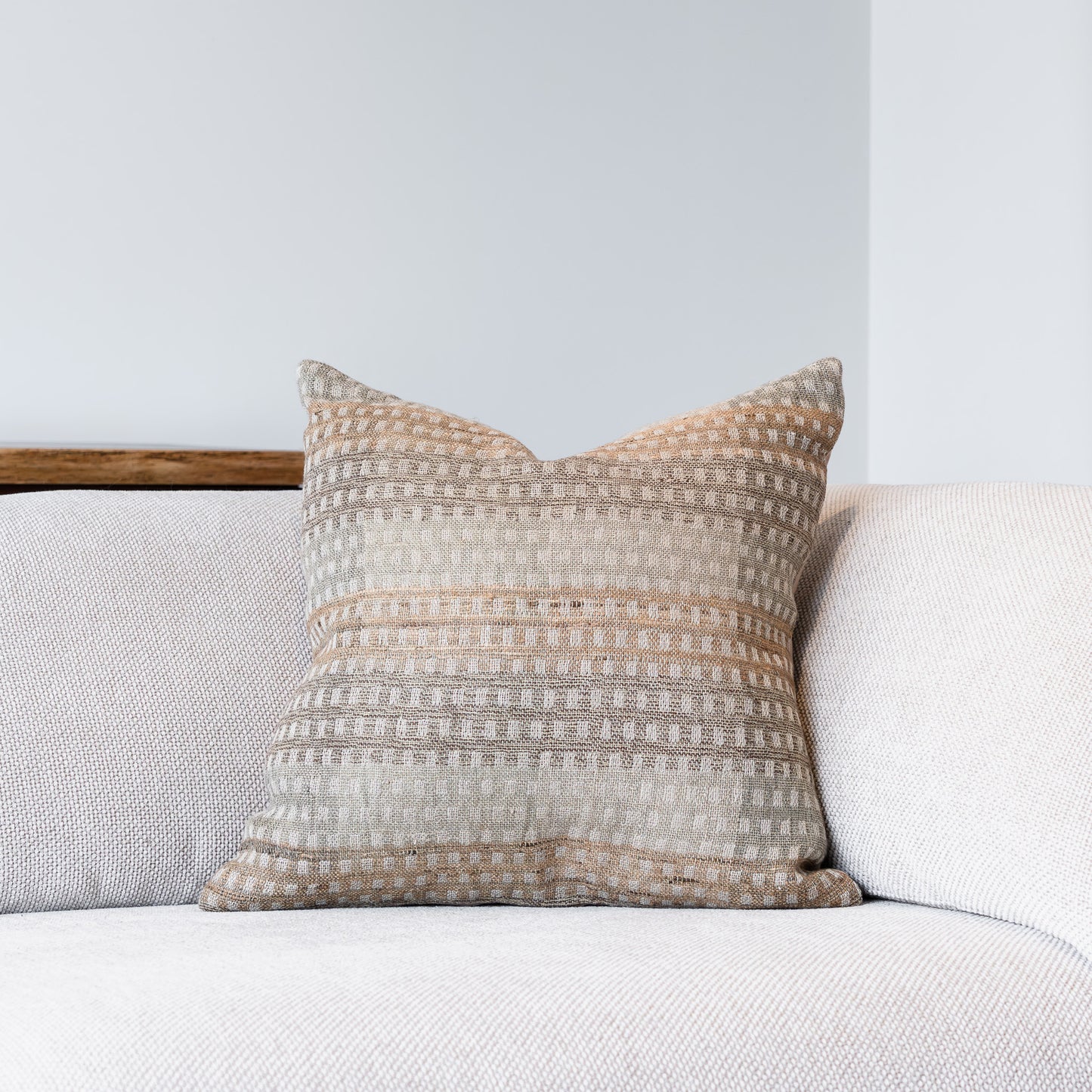 A square throw pillow with a textured, woven horizontal stripe pattern in beige, cream, and light brown, resting on a light gray sofa against a neutral wall.