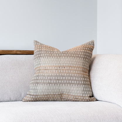 A square throw pillow with a textured, woven horizontal stripe pattern in beige, cream, and light brown, resting on a light gray sofa against a neutral wall.