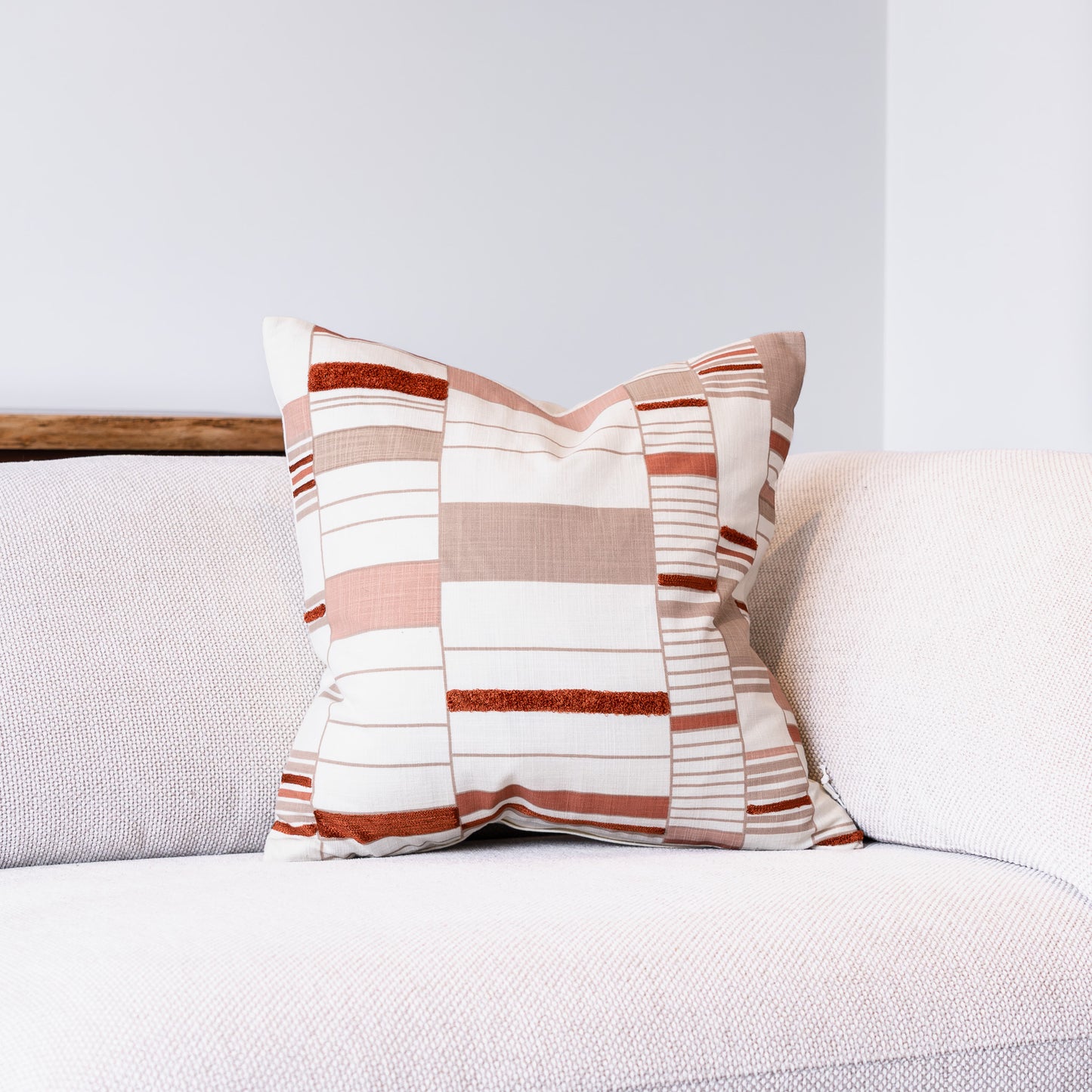 A square throw pillow with a patchwork stripe pattern in beige, white, and terracotta, resting on a light gray textured sofa.