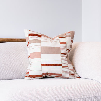 A square throw pillow with a patchwork stripe pattern in beige, white, and terracotta, resting on a light gray textured sofa.