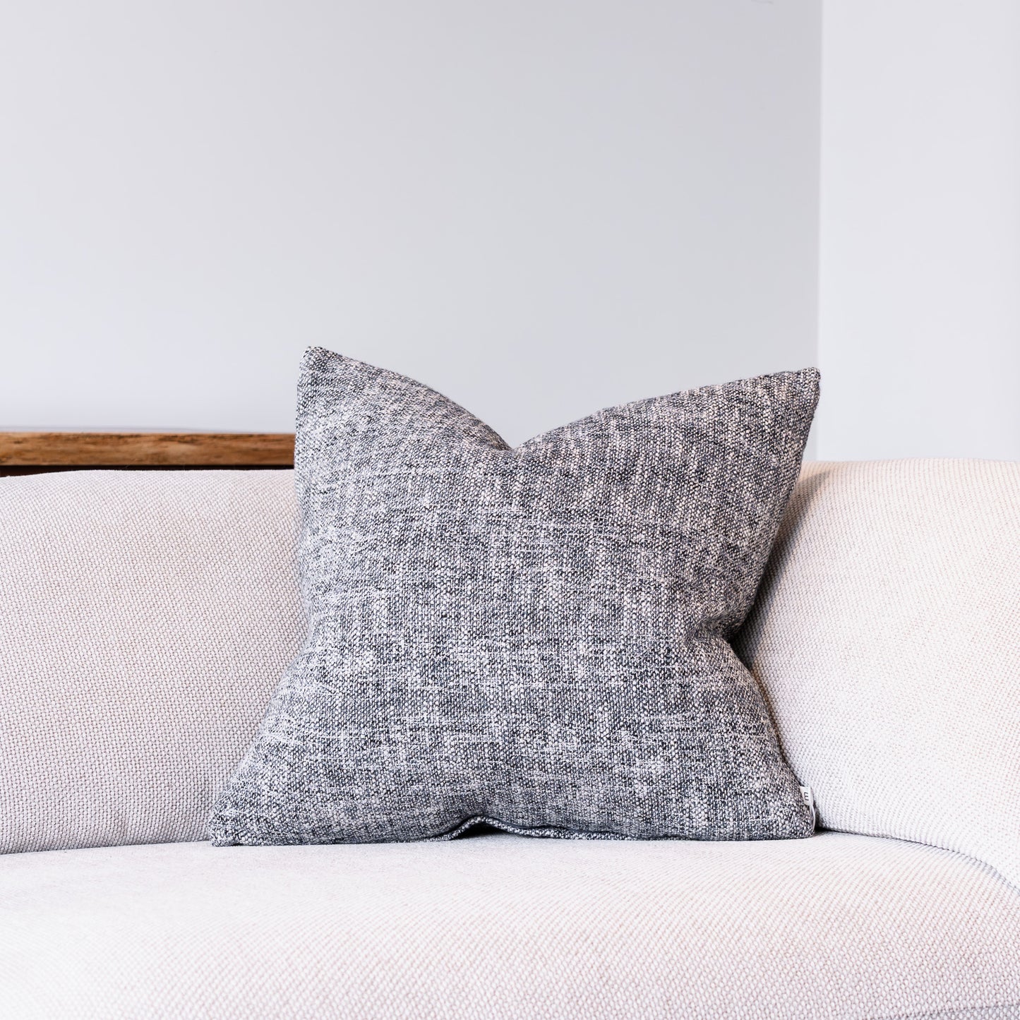 A square throw pillow with a textured gray and white woven pattern, resting on a light beige sofa against a neutral gray wall.
