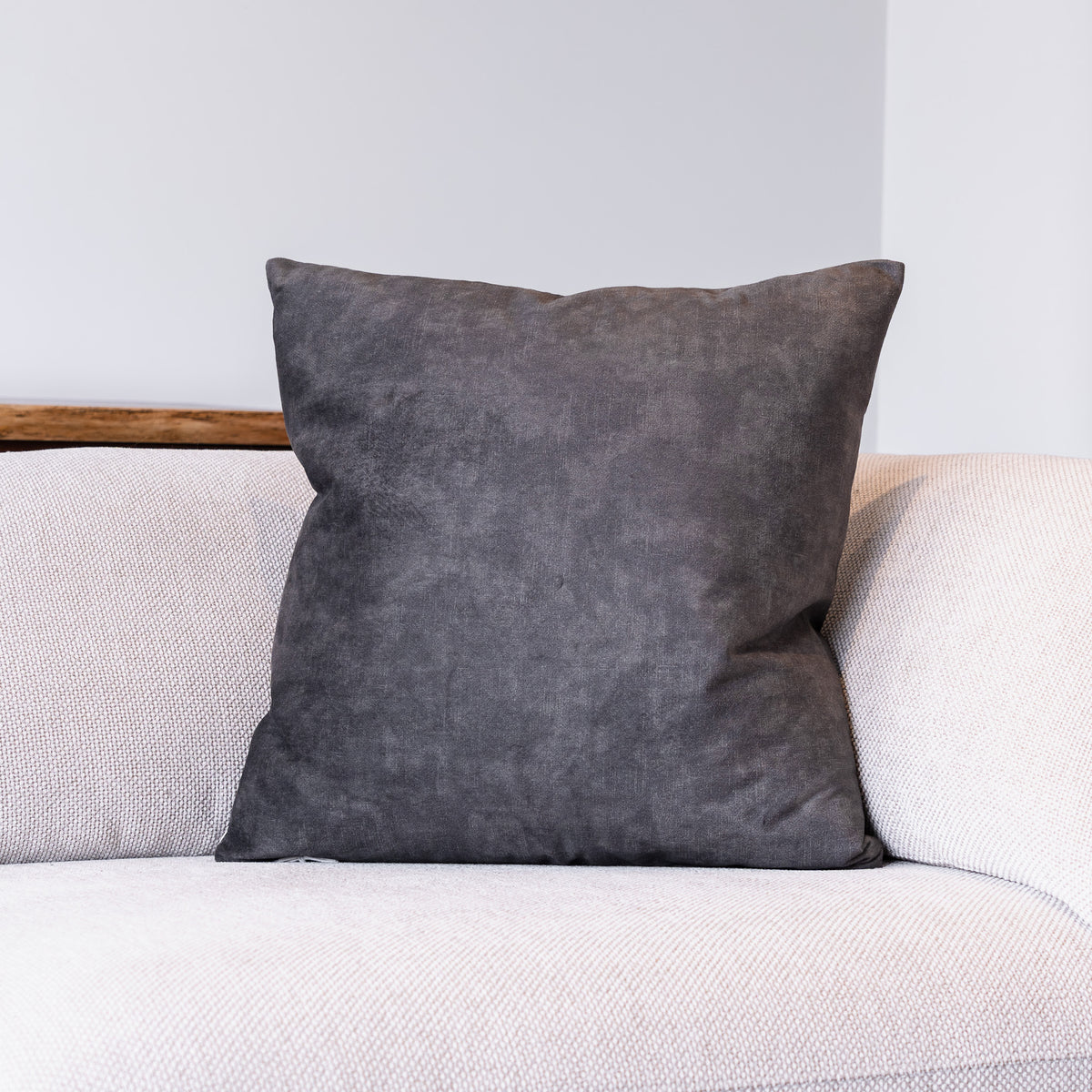 A square dark gray throw pillow with a textured suede-like finish, resting on a light beige textured sofa against a neutral gray wall.