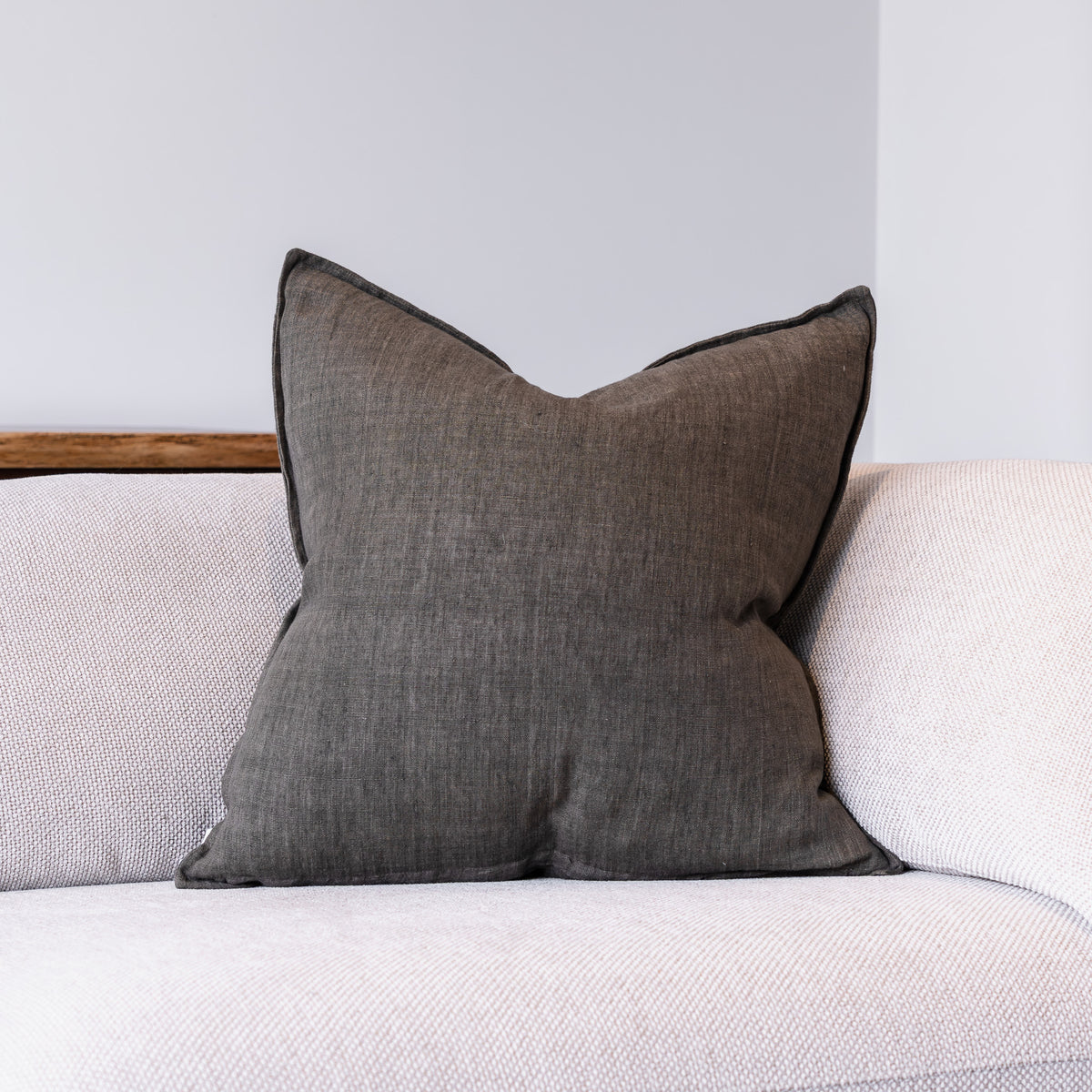 A square dark gray throw pillow with piped edges, resting on a light beige textured sofa against a neutral gray wall.