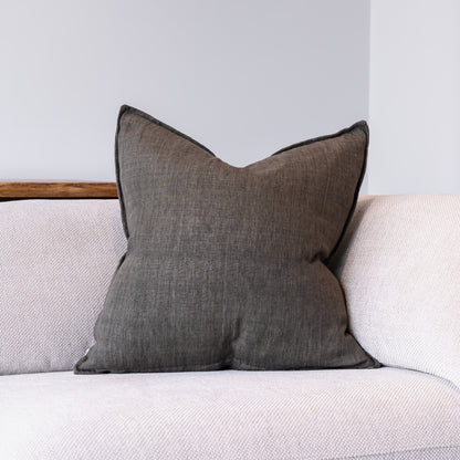 A square dark gray throw pillow with piped edges, resting on a light beige textured sofa against a neutral gray wall.