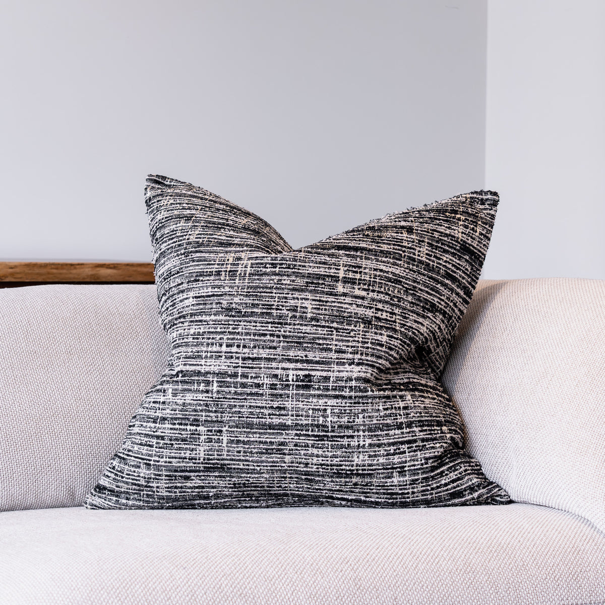 A square throw pillow with a textured black and white woven pattern, resting on a light beige sofa against a neutral gray wall.