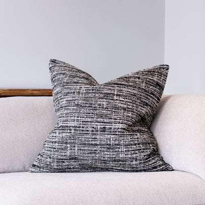 A square throw pillow with a textured black and white woven pattern, resting on a light beige sofa against a neutral gray wall.