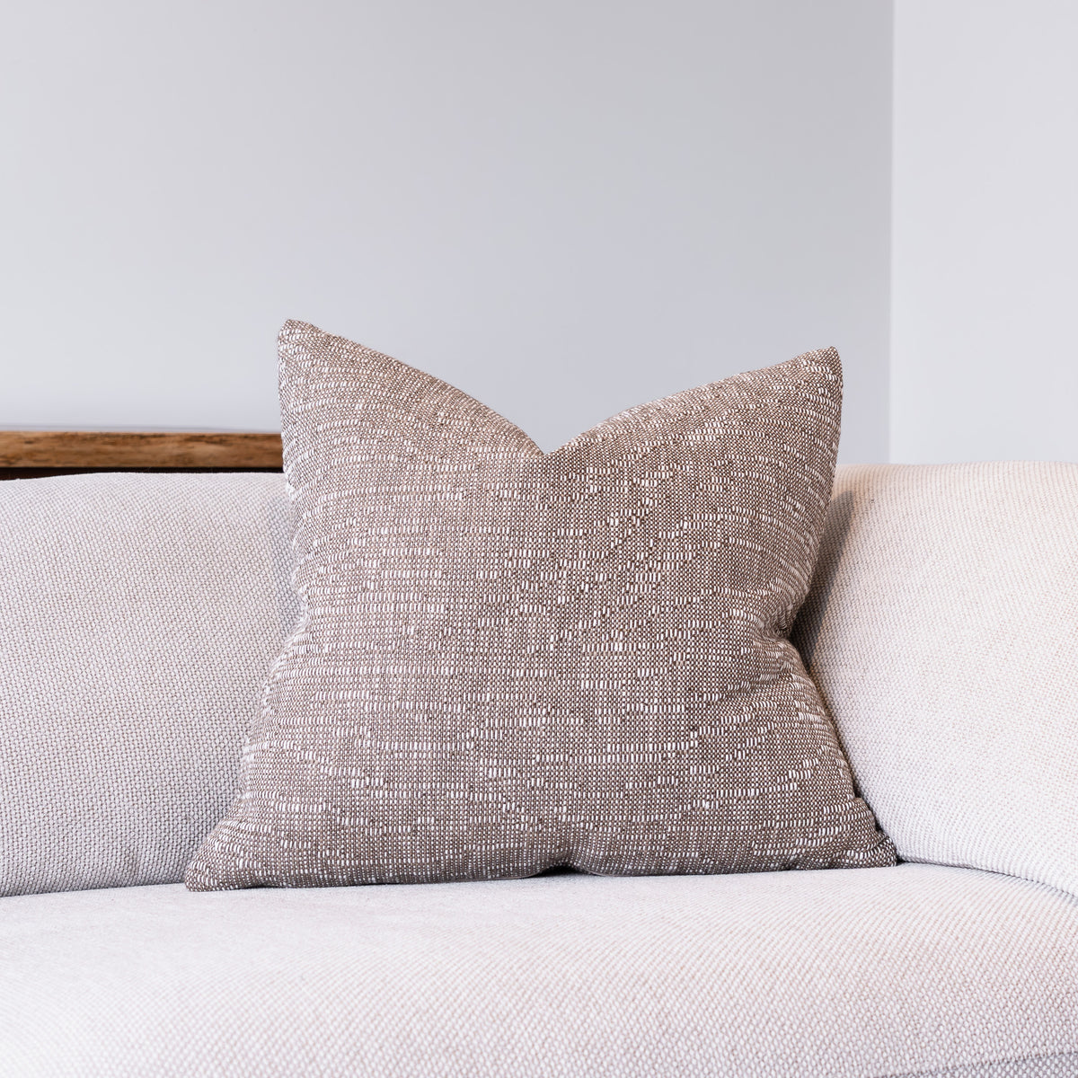 A square throw pillow with a textured, woven taupe fabric sits on a light beige sofa against a neutral gray wall.