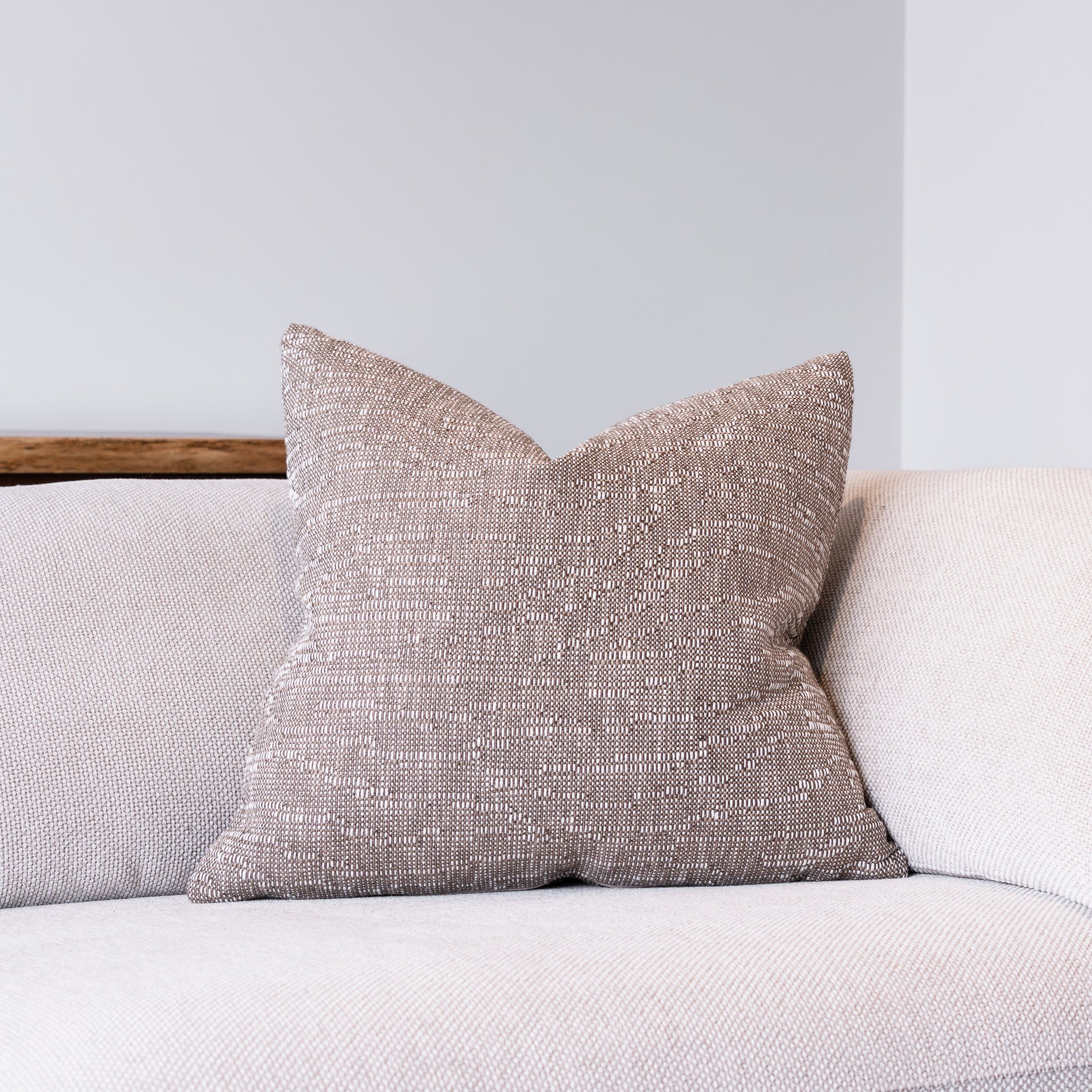A square throw pillow with a textured, woven taupe fabric sits on a light beige sofa against a neutral gray wall.