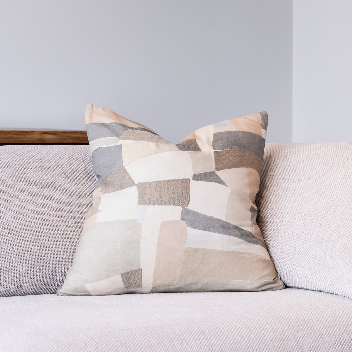 A square throw pillow with an abstract geometric patchwork pattern in beige, gray, and white, resting on a light gray textured sofa against a gray wall.