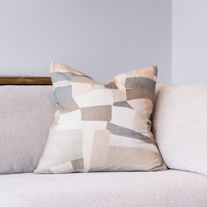 A square throw pillow with an abstract geometric patchwork pattern in beige, gray, and white, resting on a light gray textured sofa against a gray wall.
