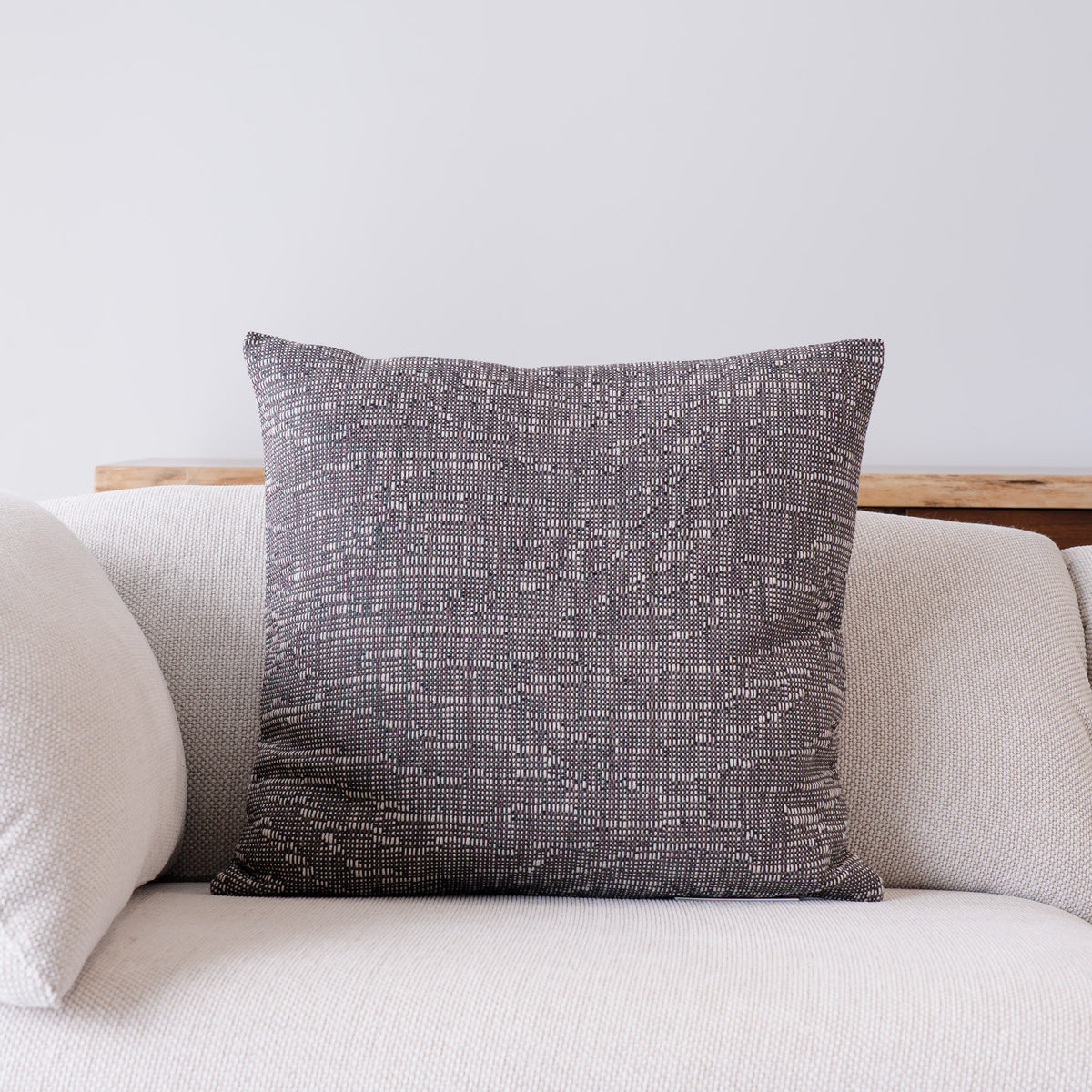 A square throw pillow with a textured, woven charcoal gray and off-white pattern, displayed on a light beige sofa against a neutral wall.
