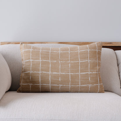 A rectangular throw pillow with a beige and white hand-drawn grid pattern, resting on a light beige textured sofa against a neutral gray wall.