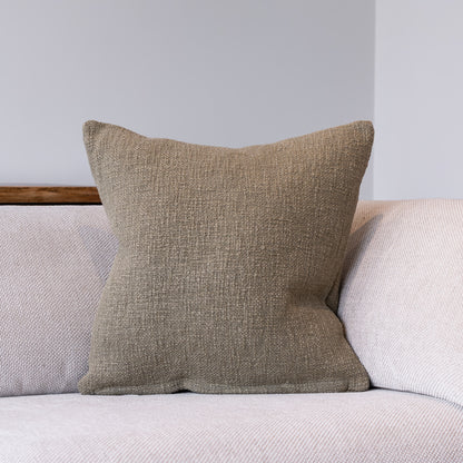 A square throw pillow in a textured taupe-brown woven fabric, resting on a light beige sofa against a neutral gray wall.