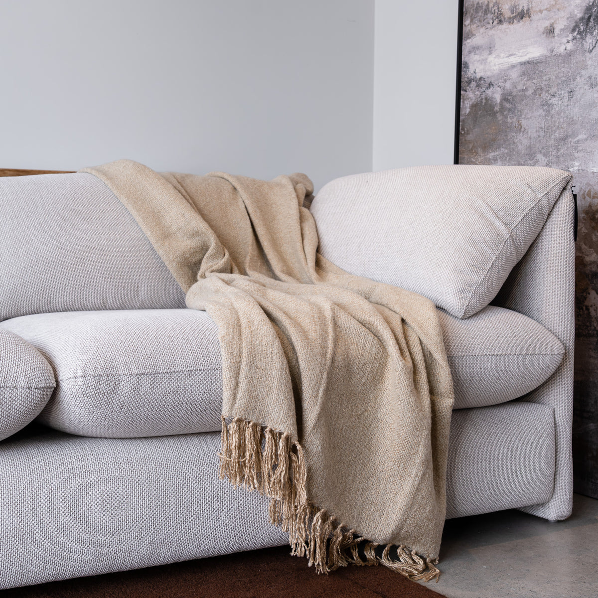 A light beige woven throw blanket with fringed edges draped over a modern light gray sofa in a minimalist room setting.