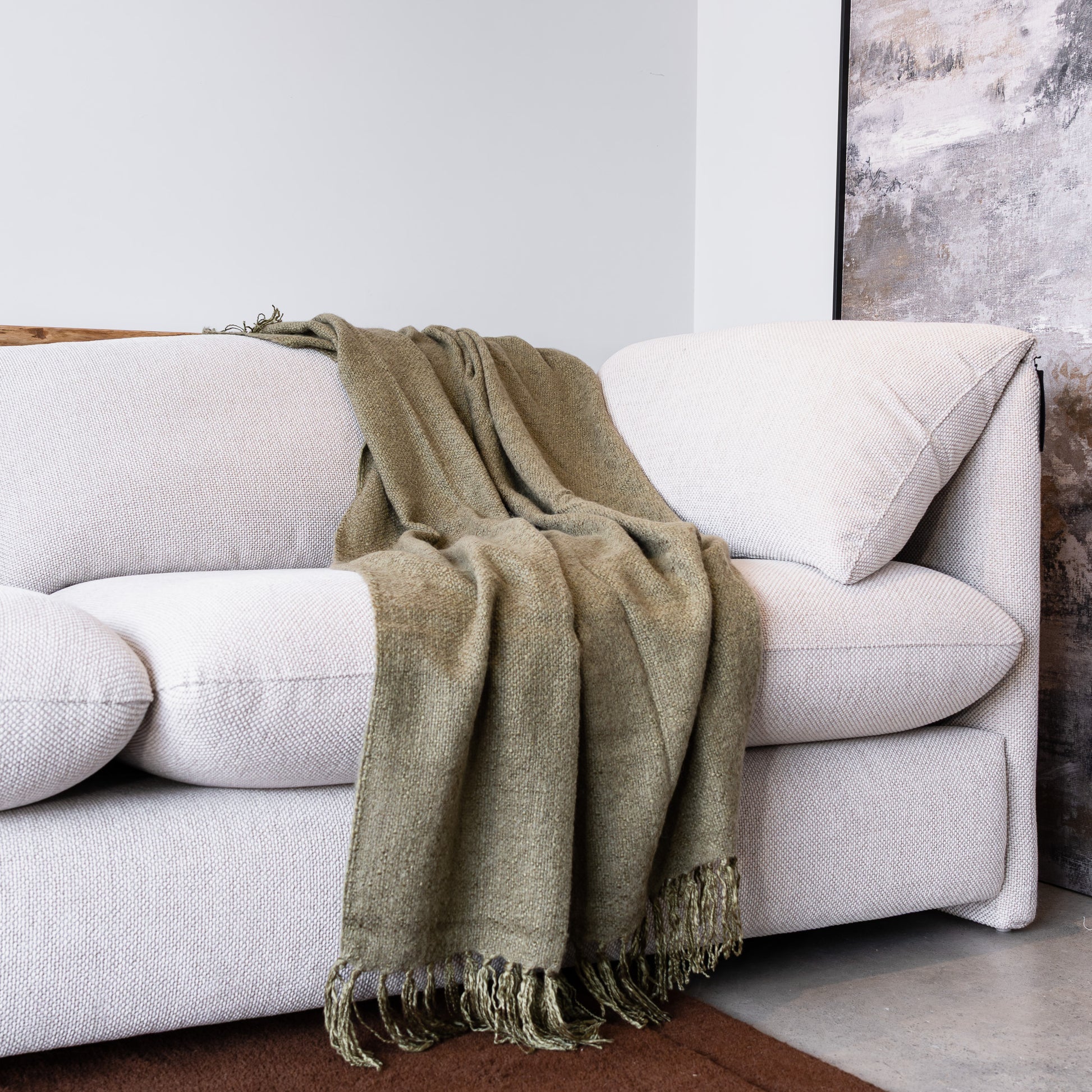 An olive green woven throw blanket with fringe detailing draped over a light beige fabric sofa in a modern living room setting.